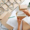 Female box maker working behind a table in a private workshop, folding, shaping a box, tools scattered across. Rulers, pencils and special board with a cutting form.