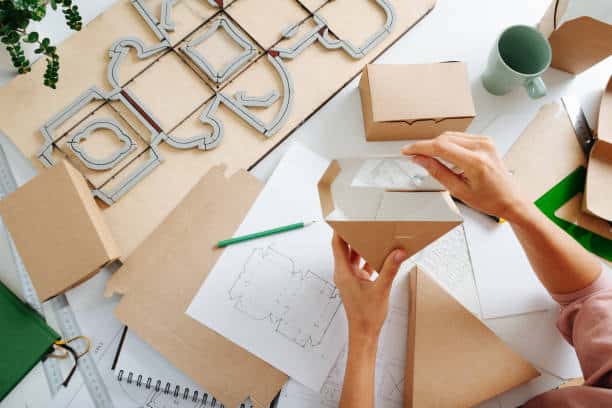 Female box maker working behind a table in a private workshop, folding, shaping a box, tools scattered across. Rulers, pencils and special board with a cutting form.