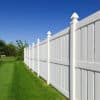 New and contemporary white vinyl fence running across a nicely landscaped backyard with lawn and blue sky in the background.