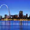 The city of St. Louis, Missouri, as seen from across the river. The Gateway Arch dominates the left upper section of the photo, more than twice as high as any of the buildings in the photo. In a horizontal line across the background and behind the Gateway Arch are downtown buildings and some skyscrapers. Lights are just coming on in the buildings even though there is still light in the sky. A line of beach lies between the buildings and the river, which is a dark blue, mirroring the dark blue of the sky.