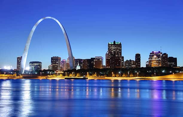 The city of St. Louis, Missouri, as seen from across the river. The Gateway Arch dominates the left upper section of the photo, more than twice as high as any of the buildings in the photo. In a horizontal line across the background and behind the Gateway Arch are downtown buildings and some skyscrapers. Lights are just coming on in the buildings even though there is still light in the sky. A line of beach lies between the buildings and the river, which is a dark blue, mirroring the dark blue of the sky.