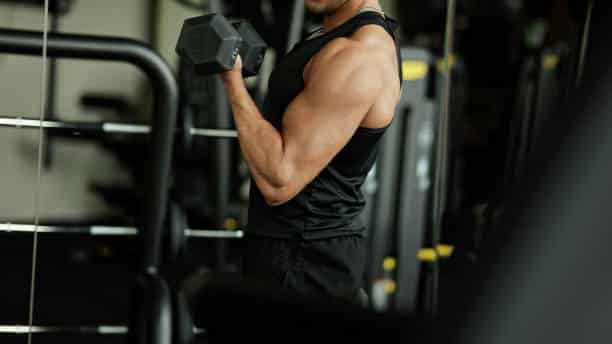 A young male athlete is captured mid-workout at the gym, intensely focused on performing bicep curls with heavy dumbbells. His posture and stance reflect a deep concentration on form and technique, ensuring maximum muscle engagement and activation throughout the exercise. The gym setting includes various fitness equipment in the background, highlighting a professional and well-equipped environment. Wearing a sleeveless black sports shirt, his well-defined arm and shoulder muscles are accentuated, showing his dedication to bodybuilding and fitness. With a determined expression, the athlete watches himself in the mirror, using his reflection to correct his form and maintain his concentration during the lift. This scene portrays the intensity and discipline required in strength training, as well as the physical strength achieved through rigorous workouts.