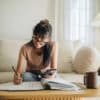 A woman sits at her living room with smartphone and financial reports doing her monthly budget.