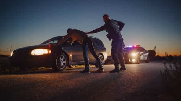 A Professional Middle Aged Policeman Performing a Pat-Down Search on a Fellon With his Hands on Car Hood. Documentary-like Shot of Procedure of Arresting Suspects. Experienced Cop Looking for Weapons