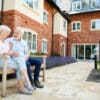 Retired Couple Sitting On Bench With Hot Drink In Assisted Living Facility