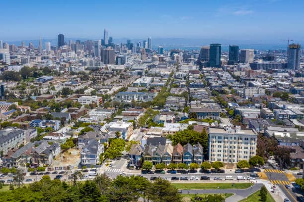 An aerial view of the colorful Row Houses referred to as "The Painted Ladies" in San Francisco. A sunny day with a clear view of the Financial District and San Francisco Bay in the background.