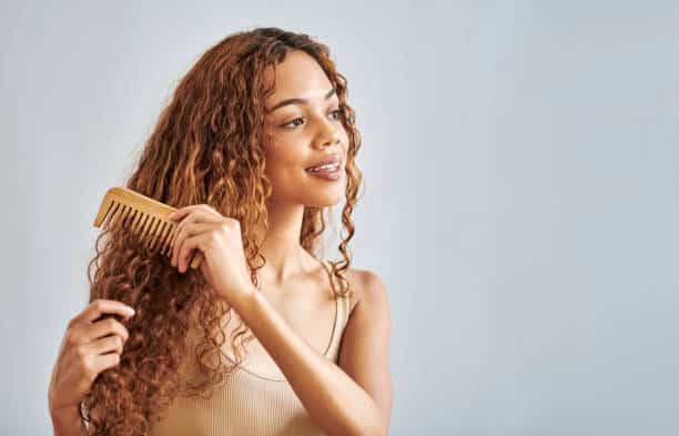 Beauty, hair and brush with a woman brushing her curly hairstyle in studio on a gray background with mockup. Face, hair care and comb with an attractive young female combing her beautiful curls