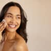 Beautiful young multiethnic woman with bare shoulder removing make up with cotton pad on cheek isolated against beige background. Smiling hispanic girl applying dry powder using cosmetic cushion on skin. Skincare and beauty routine concept.