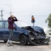 Woman with shocked face looking on her damaged car, holding head in hands. Car accident with frontal impact. Red traffic light is on background.