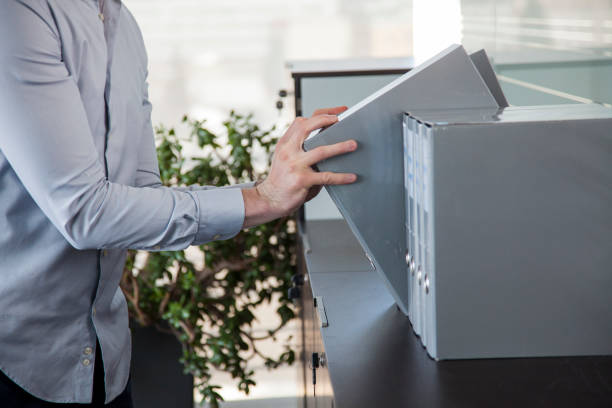 Ring binders on a shelf of a modern office with plan and window in the background. Secretary taking one.