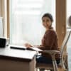 Modern day worker. Portrait of happy biracial business woman freelancer sit by computer at comfy workplace at corporate workspace or at home. Smiling young indian lady office employee look at camera