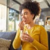 Young happy mixed race woman taking medication with water at home. One hispanic female with a curly afro taking a vitamin for good health while sitting on the couch at home. Woman drinking a supplement