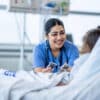 A female nurse of Middle Eastern decent sits at the edge of a hospital bed as she check in on her young patient. She is wearing blue scrubs and is attempting to cheer the young girl up.